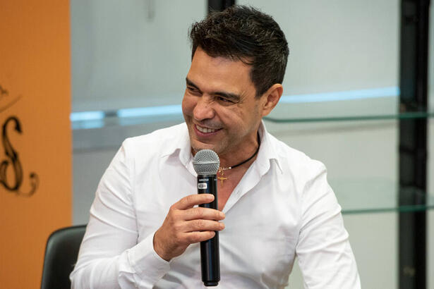 Zezé Di Camargo, com uma roupa branca e microfone na mão, em trecho de ume evento