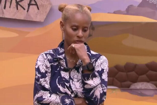 Aline Wirley com a mão no queixo, de camisa social branca com azul, cabelo preso, em pé, no Quarto Deserto, do BBB 23