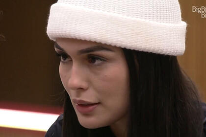 Larissa, vestindo uma toca branca, em conversa na cozinha do BBB 23. Ela olha fixamente para o canto esquerdo da tela