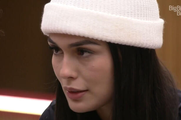 Larissa, vestindo uma toca branca, em conversa na cozinha do BBB 23. Ela olha fixamente para o canto esquerdo da tela