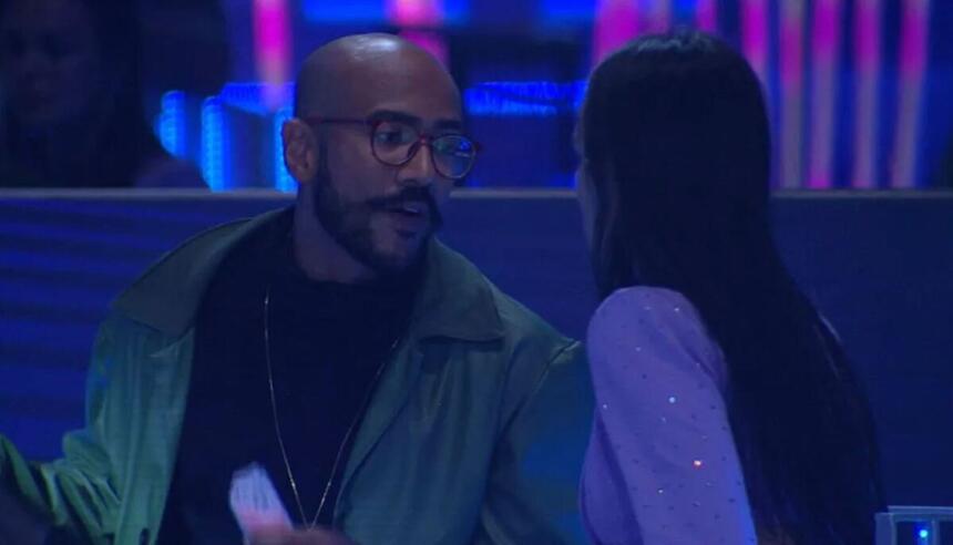 Ricardo com semblante sério de frente para Larissa que aparece de costas na imagem.