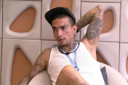 MC Guimê de camisa reggata branca, chapéu preto, colar azul sentado na varanda do BBB 23