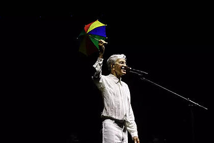 Caetano Veloso de camisa e calça branca suspendendo uma sobrinha de frevo durante show em recife
