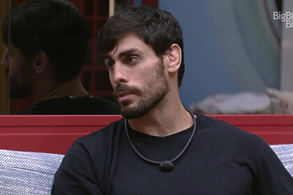 Cara de Sapato sentado na sala do BBB 23, vestindo uma camiseta preta e olhando para o lado
