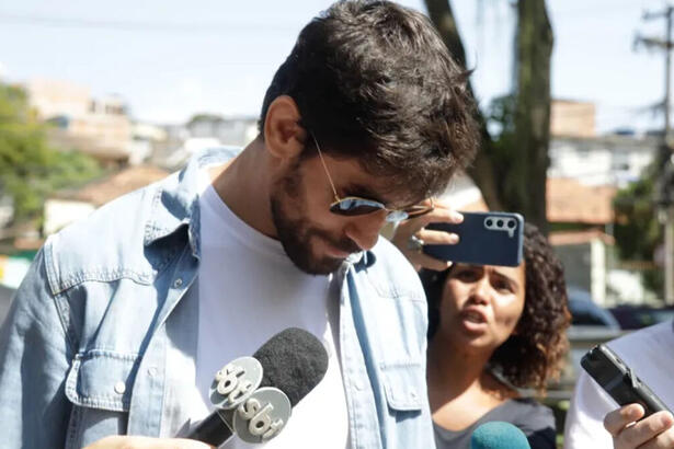 Cara de Sapato, de óculos escuros e cabeça baixa, rodeado de microfones de jornalistas