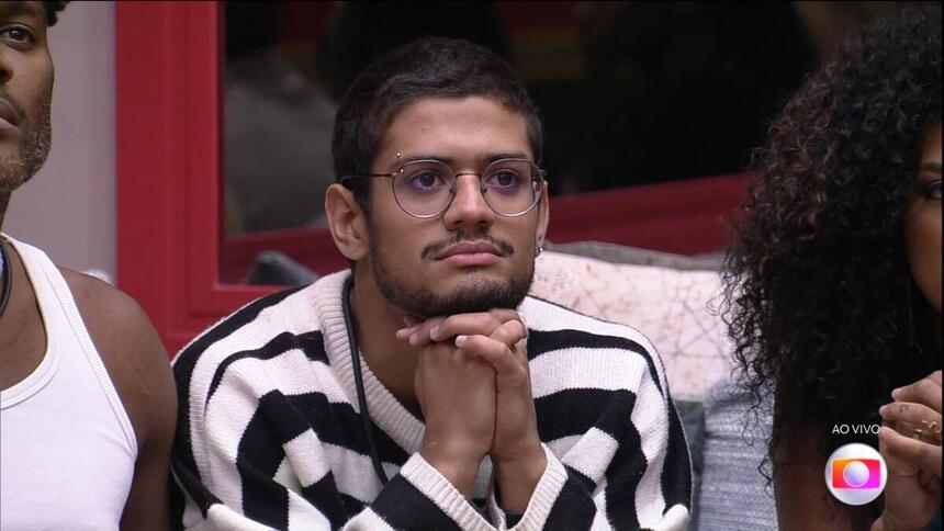 Gabriel Santana com camisa manga longa listrada em branco e preto, com as mãos dadas, semblante sério, oculos, sentado no sofá