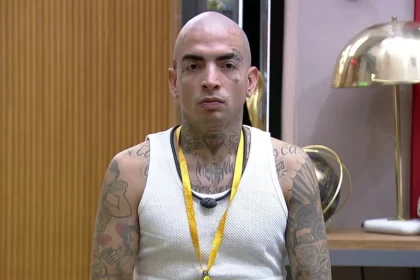 MC Guimê careca, de camisa regata branca, se olhando no espelho