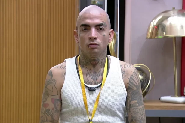 MC Guimê careca, de camisa regata branca, se olhando no espelho