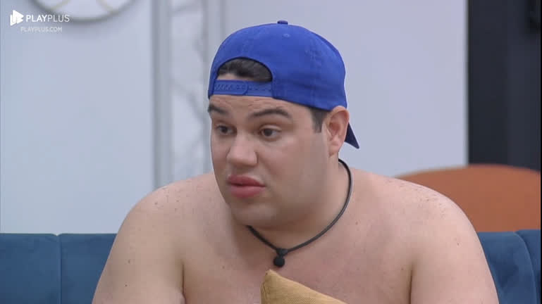 Erick Ricarte de boné azul, sem camisa, sentado no sofá