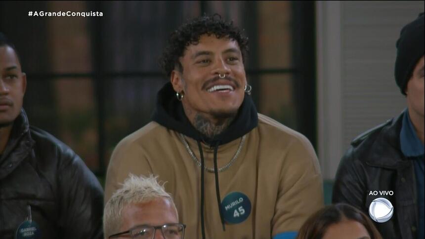 Murilo de camisa bege, sorrindo, comemorando o resultado da votação de A Grande Conquista