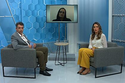Renato Dias sentado nos estúdios do Aparecida Debate, com um dos convidados sentado a sua frente e outro através de videoconferecia, pela televisão no centro do estúdio