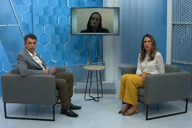 Renato Dias sentado nos estúdios do Aparecida Debate, com um dos convidados sentado a sua frente e outro através de videoconferecia, pela televisão no centro do estúdio