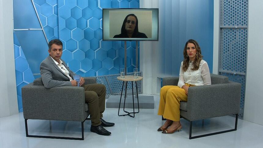Renato Dias sentado nos estúdios do Aparecida Debate, com um dos convidados sentado a sua frente e outro através de videoconferecia, pela televisão no centro do estúdio