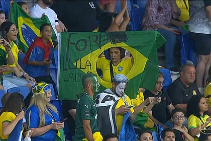 Mulher segura bandeira com frase Lula Ladrão em transmissão do amistoso Brasil x Guiné na TV Globo