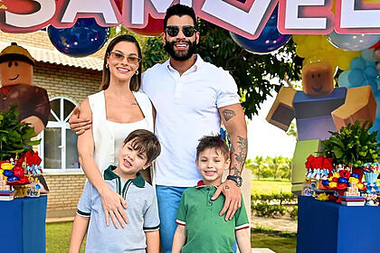 Gusttavo Lima de oculos de sol, sorrindo, de camisa branca, ao lado da esposa Andressa Suita e os filhos durante festa.