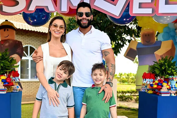 Gusttavo Lima de oculos de sol, sorrindo, de camisa branca, ao lado da esposa Andressa Suita e os filhos durante festa.
