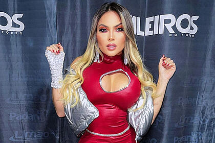 Kally Fonseca com semblante sério, braços levantados, com look prata e vermelho, durante bastidores de show