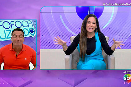Leo dias de camisa polo vermelha, sentado numa bancada, nos estúdios do fofocalizando; isabele benito de camisa preta e macacãoa azul, sentada, de cabelo solto, gesticulando, nos estúdios do sbt