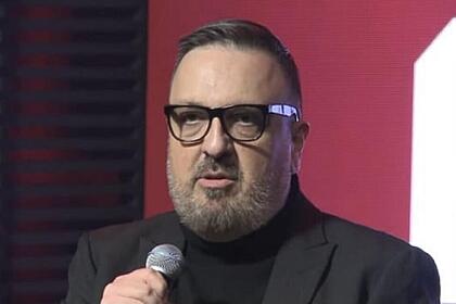 Rodrigo Carelli com semblante sério, usando óculos de grau, palitó e camisa preta, segurando um microfone durante coletiva