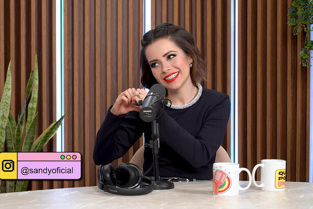 Sandy sorrindo, fazendo coração com as mãos, sentada durantre entrevista no podcast