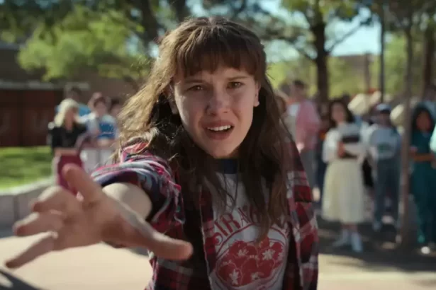 personagem de stranger things de camisa em estampa preta, vermelha e branca, com o braço pra frente, durante gravação de série