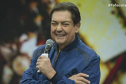 Faustão sorrindo e segurando um microfone no cenário do seu programa da Band