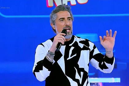 Marcos Mion apresentando o Caldeirão. Ele está de camisa preto e branco e com o braço esquerdo levantado