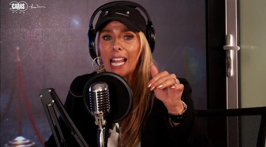 Adriane Galisteu de chapéu e camisa pretos, sentada, com fone de ouvido, durante entrevista no Podcaras