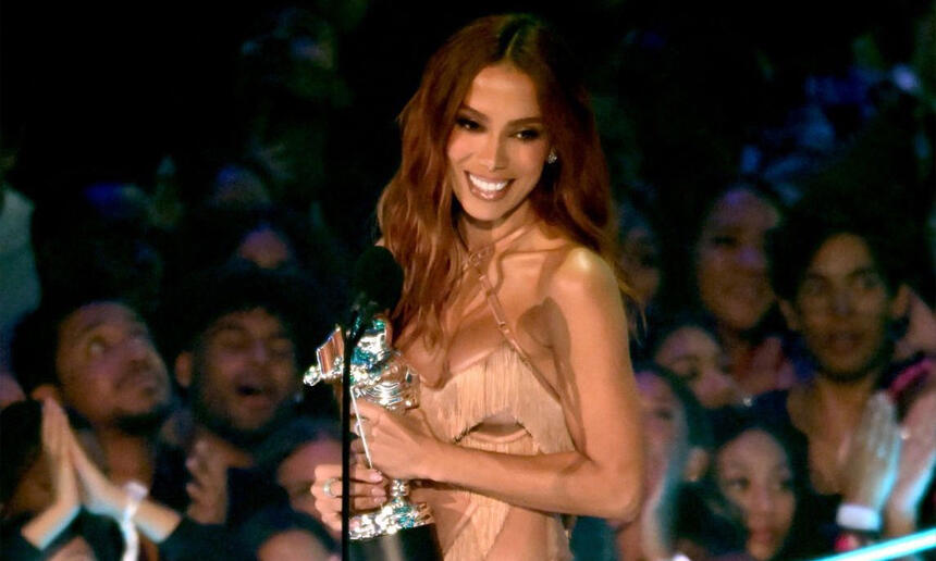Anitta sorrindo, de vestido dourado, em pé, segurando o prêmio do VMA 2023