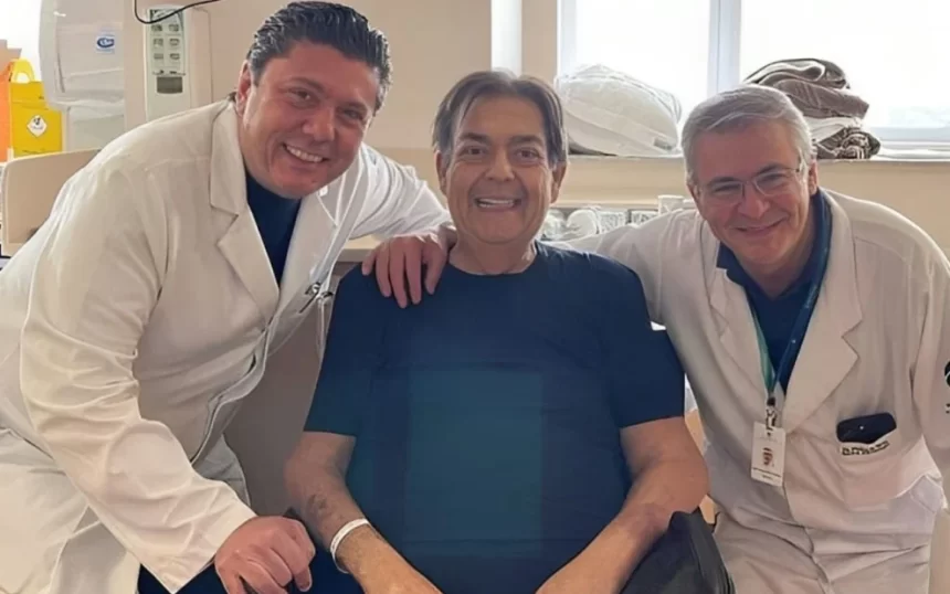 Faustão de camisa azul, sentado numa cadeira de roda, sorrindo, ao lado de médicos em um hospital