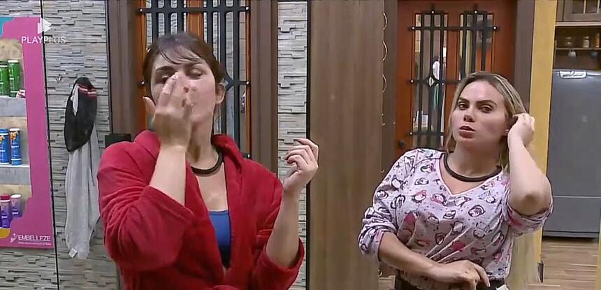 Nadja com a mão no rosto passando um creme do lado de Kally com semblante sério com a mão na orelha