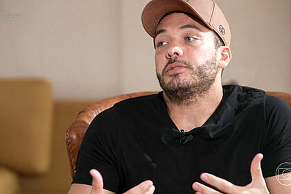 Wesley Safadão de boné marrom, camisa preta, sentado num sofá marrom, durante gravação de entrevista para o Fantástico, da TV Globo