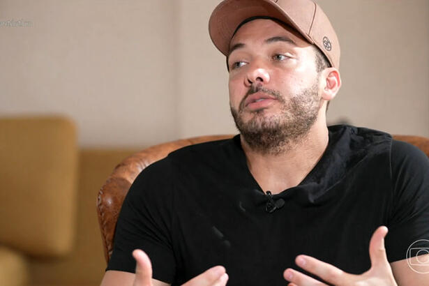 Wesley Safadão de boné marrom, camisa preta, sentado num sofá marrom, durante gravação de entrevista para o Fantástico, da TV Globo