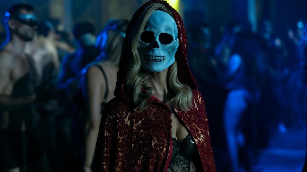 Mulher loira com máscara de caveira vestindo uma capa de renda vermelha em uma festa.