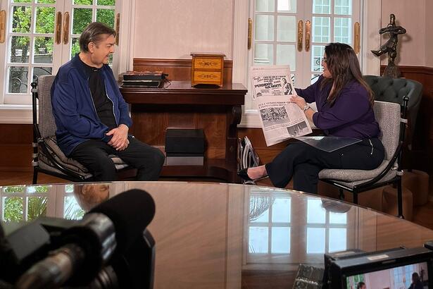 Faustão sorrindo enquanto Fabíola Reipert lendo um jornal antigo durante gravação de entrevista para o Domingo Espetacular