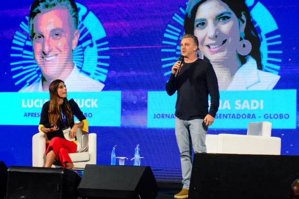 Luciano Huck em pé, de camisa preta manga longa, calça jeans, no palco da Rio Innovation Week