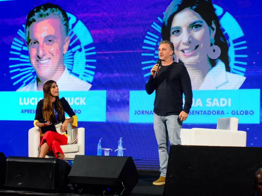 Luciano Huck em pé, de camisa preta manga longa, calça jeans, no palco da Rio Innovation Week