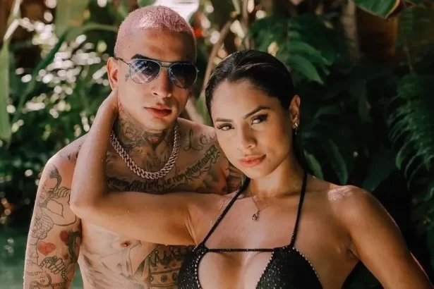 MC Guimê sem camisa, de oculos de sol, com semblante sério, abraçado com Lexa, que está de biquini preto e cabelo preso, molhado