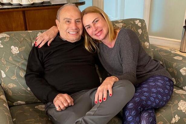 Marilene Saade de camisa mangalonga cinza, sentada num sofá, de calça azul, sorrindo, ao lado de Stênio Garcia