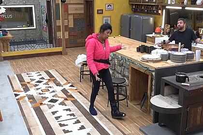 Marcia Fu de uma lado da cozinha em Pé, com uma blusa rosa escura, apontado o dedo para Sander, que se encontra do outro lado perto do fogão.