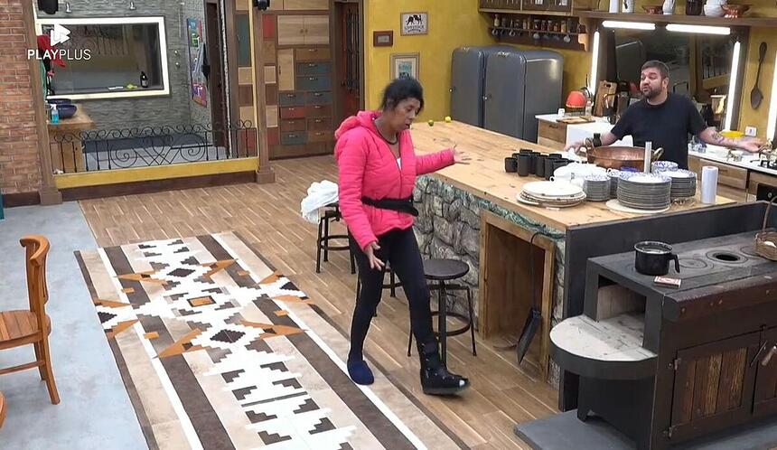 Marcia Fu de uma lado da cozinha em Pé, com uma blusa rosa escura, apontado o dedo para Sander, que se encontra do outro lado perto do fogão.