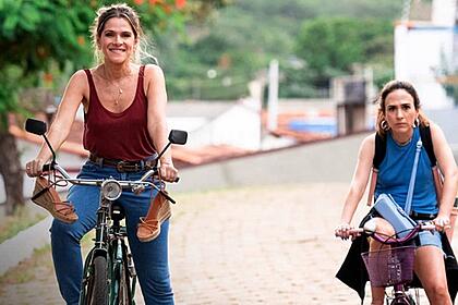 Ingrid Guimarães sorrindo, de camisa regata vermelha, calça jeans, segurando numa bicicleta parada numa praça; Imagem 2: Tatá Werneck de camisa regata, jaqueta jeans amarrada, encostada na bicicleta