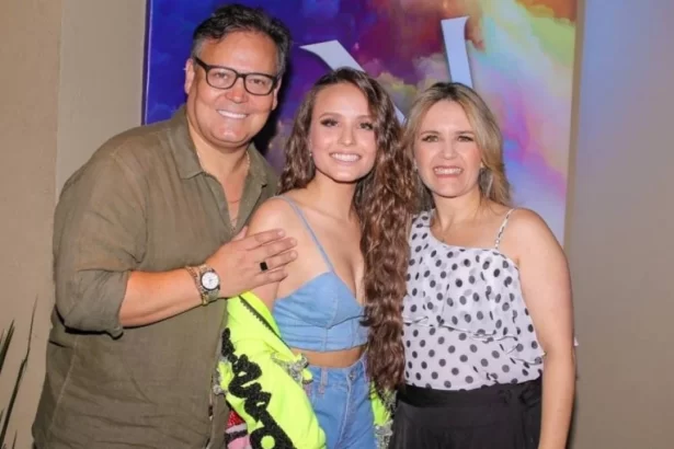 Larissa Manoela sorrindo, de cabelo solto, com o casaco aberto, ao lado dos pais