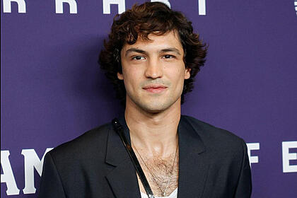 Gabriel Leone de camisa branca e blazer preto em sobreposição, cabelo preto, mais cheio, com semblante feliz em coletiva de imprensa