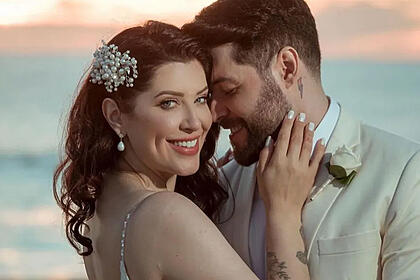 Andressa Ganacin sorrindo, vestida de noiva, com a mão no rosto de nascer num cenário paradisiaco