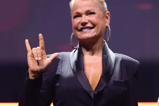 Xuxa de blazer preto, sorrindo, fazendo um simbolo com as mãos