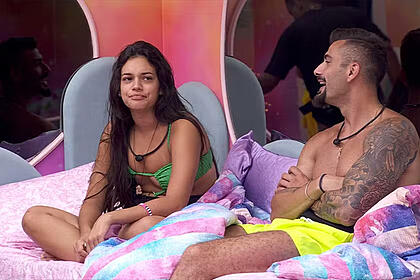Alane de cabelo solto, com as pernas cruzadas, sentada na cama ao lado de NIzam