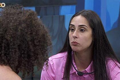 Deniziane de camisa rosa, cabelo solto, com semblante emburrado, sentada conversando com Maycon