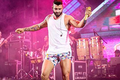 Gusttavo Lima de short curto, camisa regata branca, cantando em palco