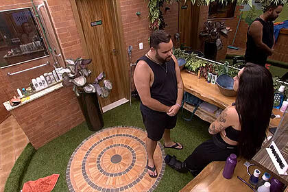 Michel de camisa e short brancos, em pé no banheiro na frente de Giovanna